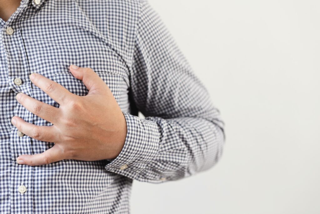 Una persona que viste una camisa a cuadros se agarra el pecho con una mano, lo que posiblemente indica incomodidad o dolor. El fondo es liso y de color blanquecino.