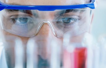 Primer plano de un científico con gafas de seguridad, observando atentamente tubos de ensayo con líquidos coloridos en un entorno de laboratorio, como si analizara datos para el informe de salud del Boletín Médico EuroEspes.